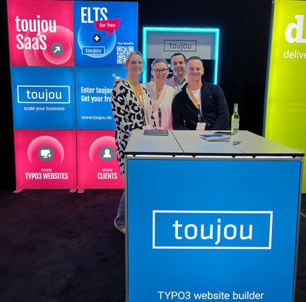 Hinter einem bunten Stand stehen vier Personen, die alle lächeln. Hinter ihnen sind weitere bunte Standwände zu sehen vor einem schwarzen Hintergrund.