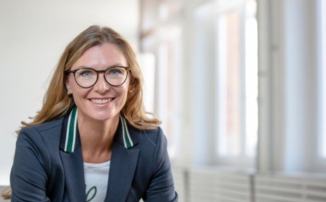 Porträtbild einer Frau mit blonden Haaren, Brille und einem dunklen Blazer