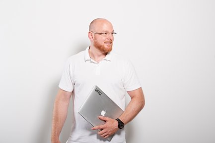 Ein Mann mit einem Bart und einer Brille lächelt, während er einen Laptop in der Hand hält, vor einem weißen Hintergrund.