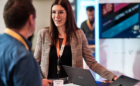 Eine Frau mit Brille und kariertem Blazer spricht mit einem Mann, während sie an einem Tisch mit Laptops stehen.