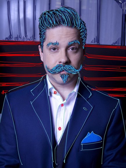 Ein Mann mit einem markanten Schnurrbart und gestyltem Haar trägt einen eleganten Anzug mit einer blauen Tasche.