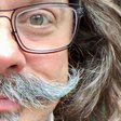 Ein Mann mit einer auffälligen, grauen Schnurrbartfrisur und Brille blickt direkt in die Kamera, seine Augen strahlen Neugier aus.