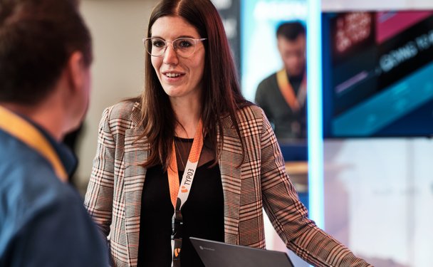 Eine Frau mit Brille und kariertem Blazer spricht mit einem Mann, während sie an einem Tisch mit Laptops stehen.