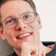 Ein junger Mann mit Brille lächelt und seine Hand als Faust vor dem Kinn