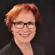 Eine lächelnde Frau mit rotem Haar und Brille trägt einen schwarzen Blazer und posiert vor einem dunklen Hintergrund.