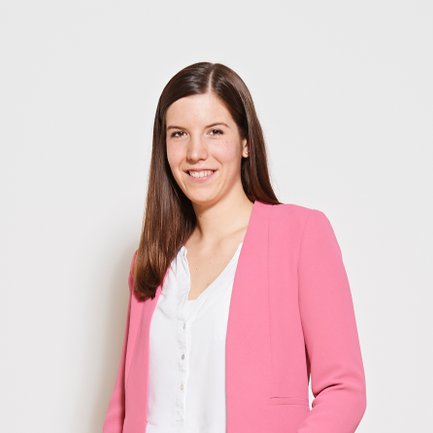 Eine junge Frau mit langen, glatten Haaren lächelt freundlich in einem pinken Blazer vor einem neutralen Hintergrund.