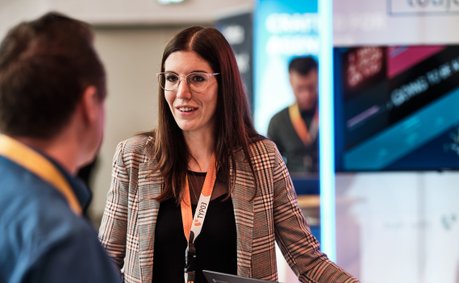 Eine Frau mit Brille und kariertem Blazer spricht mit einem Mann, während sie an einem Tisch mit Laptops stehen.