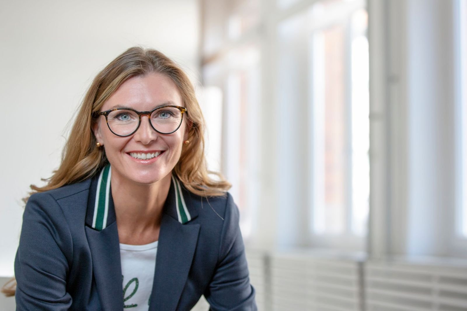 Porträtbild einer Frau mit blonden Haaren, Brille und einem dunklen Blazer
