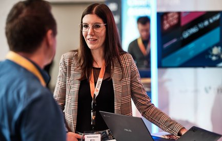 Eine Frau mit Brille und kariertem Blazer spricht mit einem Mann, während sie an einem Tisch mit Laptops stehen.