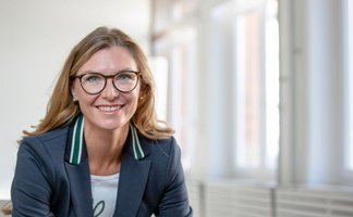 Porträtbild einer Frau mit blonden Haaren, Brille und einem dunklen Blazer