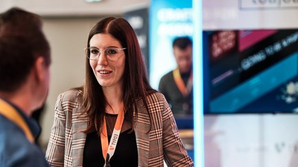 Eine Frau mit Brille und kariertem Blazer spricht mit einem Mann, während sie an einem Tisch mit Laptops stehen.